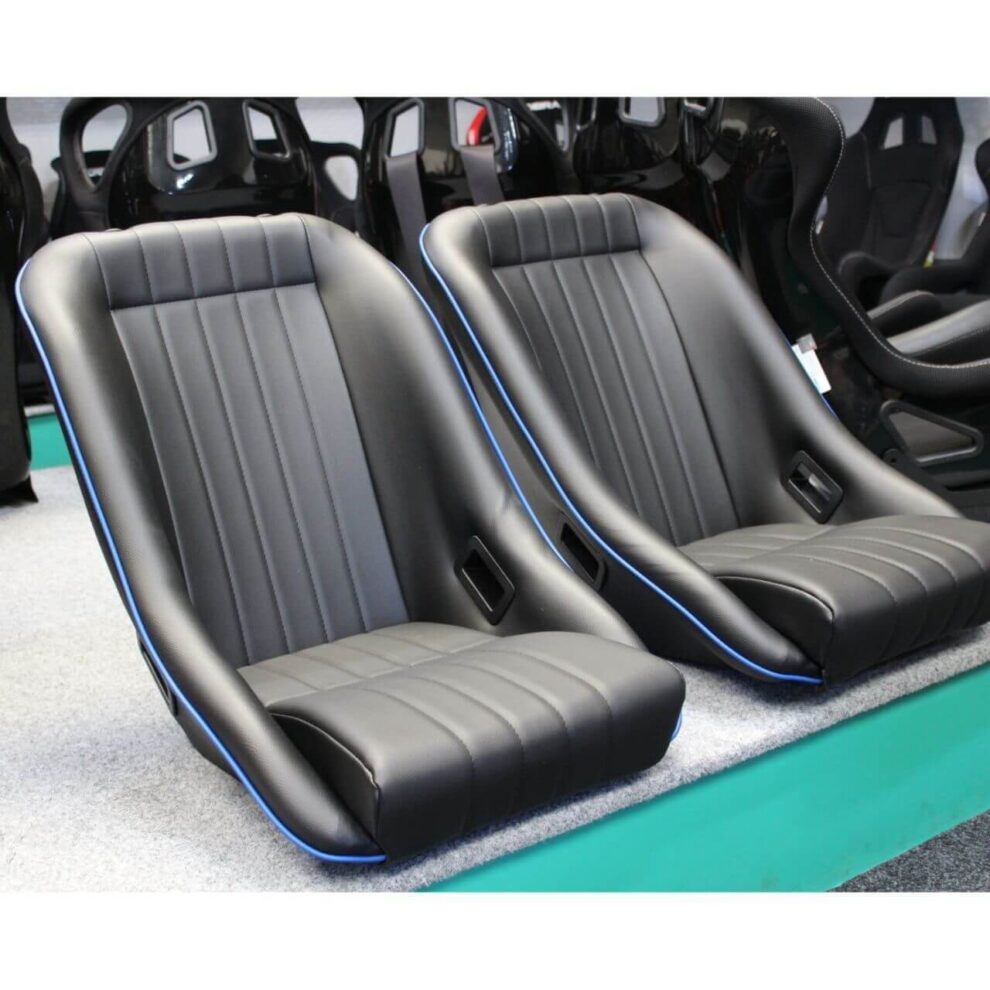 Two black classic style bucket seats with blue piping on the green showroom plinth, with FIA bucket seats on the wall behind