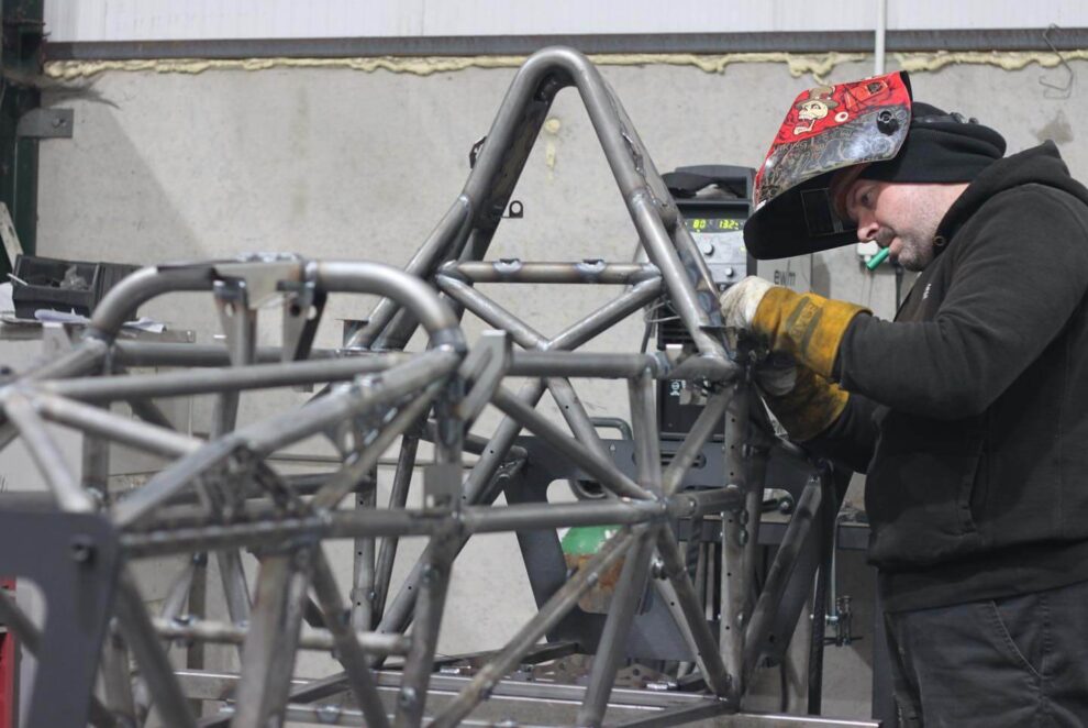 A fabricator in a welding helmet working on a bare steel tube frame in a workshop