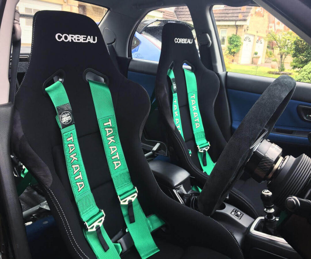 Two black Corbeau bucket seats with green Takata four point harnesses fitted in a car with a blue interior
