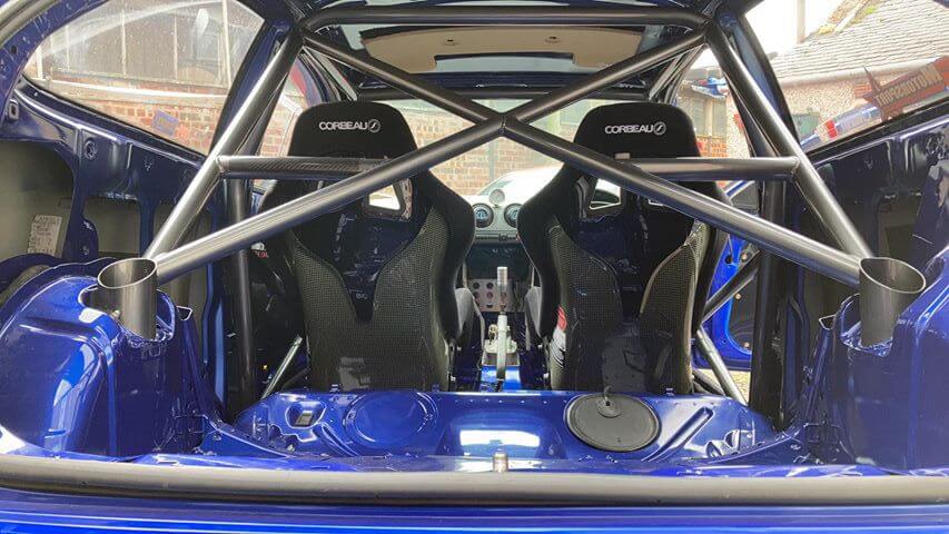 A pair of Corbeau Pro Series System 3 bucket seats seen from behind through the roll cage of a blue customer car, the woven aramid shell backs visible on both