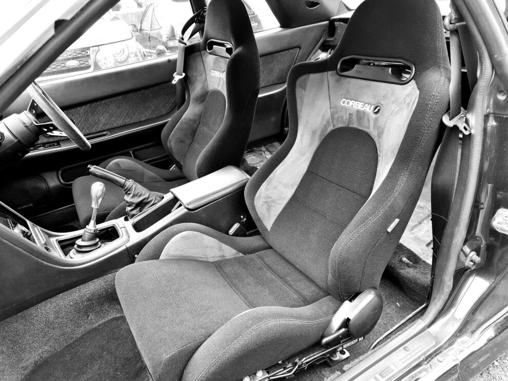 Black and white photo of two Corbeau reclining sport seats with suede panels in a Nissan Skyline R32, seen through the open passenger door