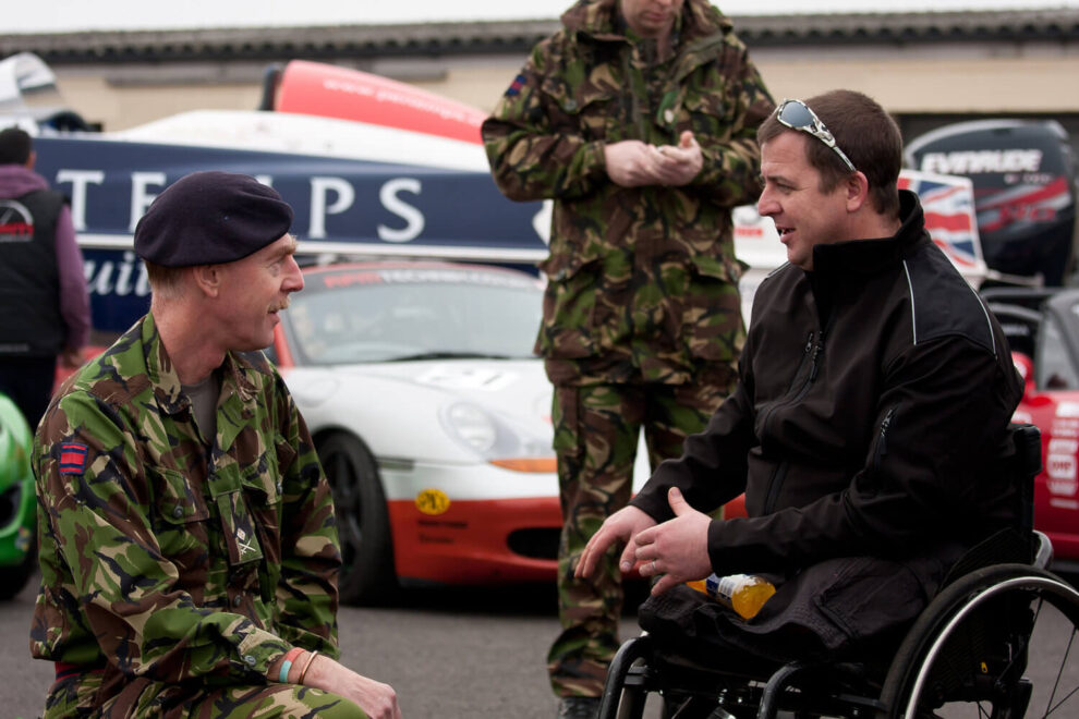Service personnel in camouflage talking with a man in a wheelchair in a race paddock, with race cars behind