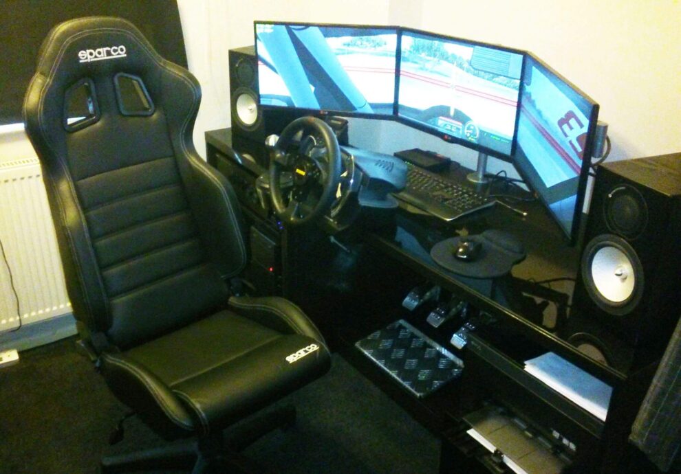 A black Sparco reclining seat on an office chair base beside a sim racing desk with a wheel, pedals and three screens