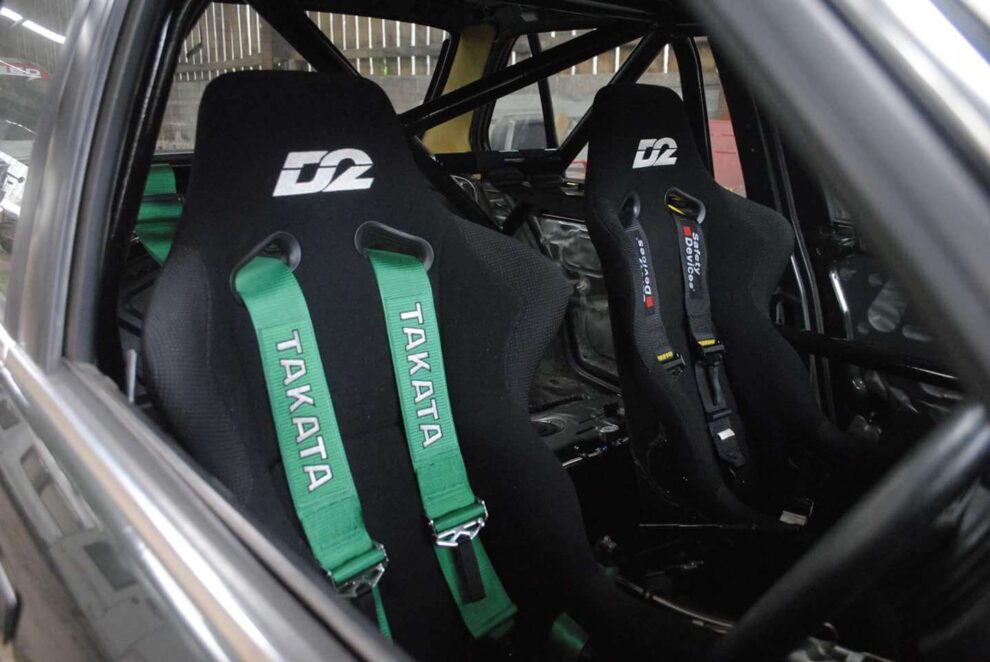 A green Takata harness on a black D2 bucket seat in a caged car, with a second seat and harness beside it