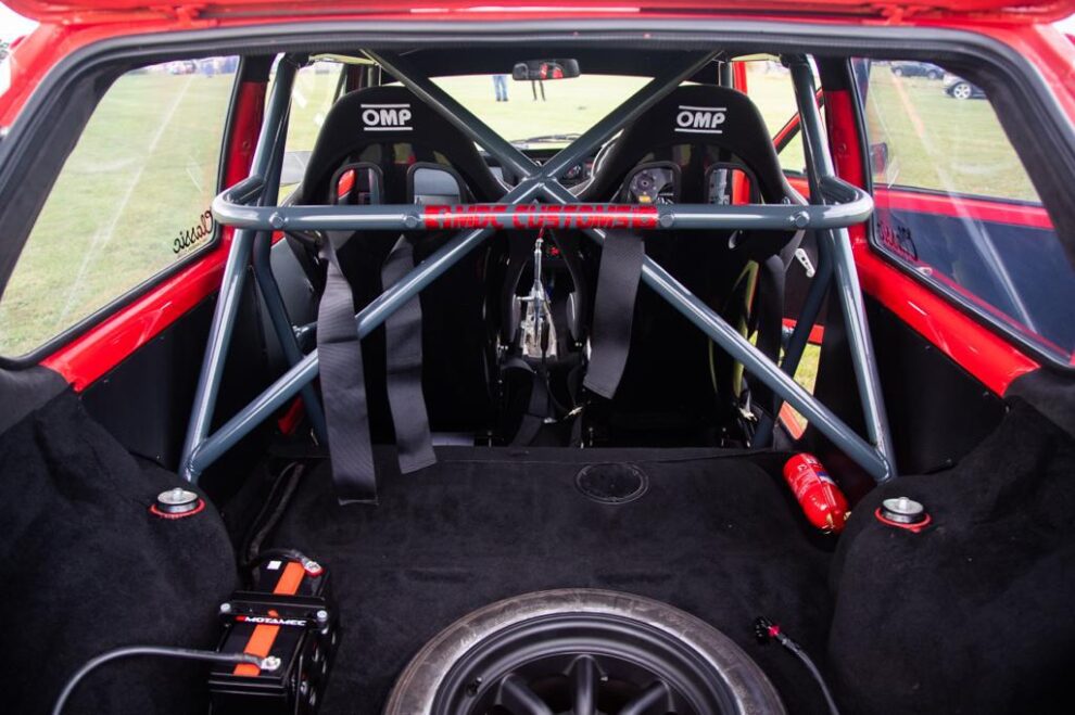 View through the tailgate of the red VW Golf showing the grey roll cage, harness straps and the backs of two OMP seats