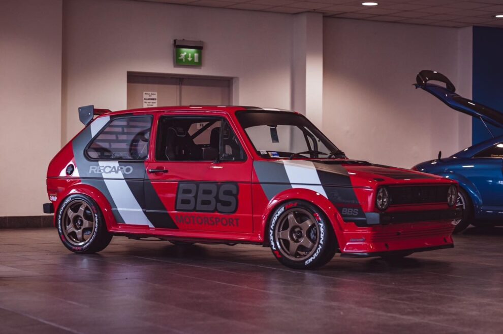 A red, white and grey liveried VW Golf Mk1 track car with gold wheels parked indoors at a show