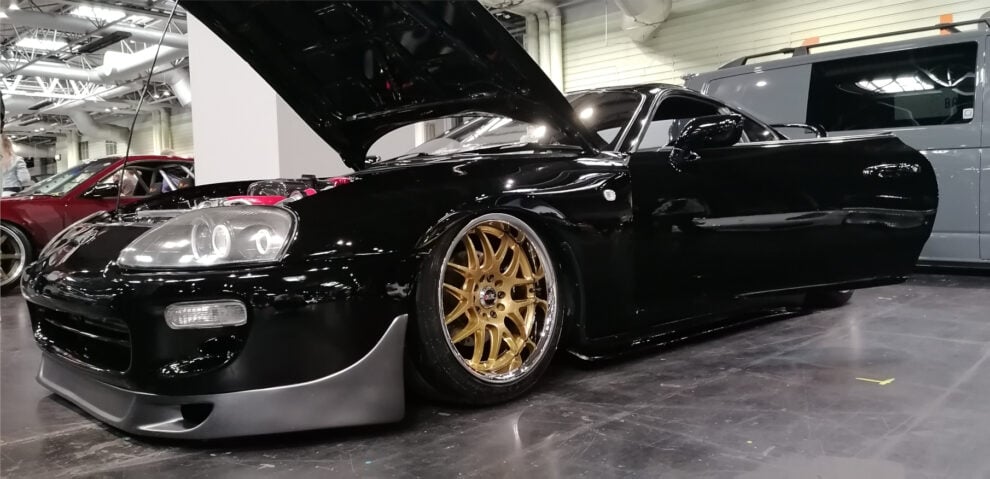 A black Toyota Supra with gold multi-spoke wheels and its bonnet raised, sitting low on the show floor inside the NEC