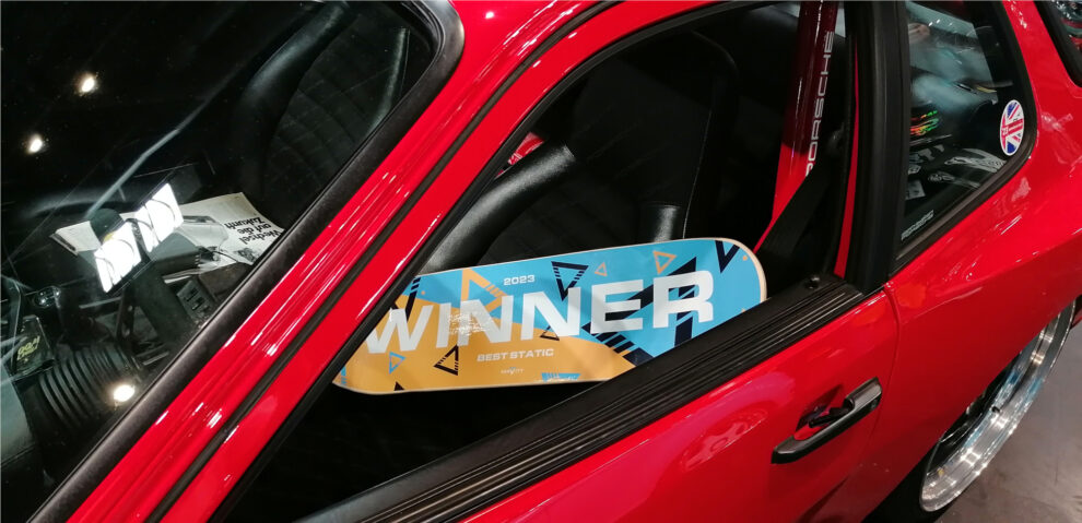 A Gravity 2023 winner board for Best Static resting inside the window of a red Porsche at the show