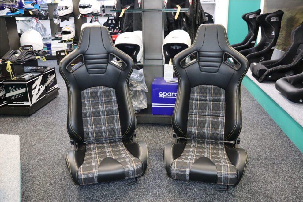 A pair of Auto Style GT reclining seats with black vinyl sides and tartan centres on the showroom floor, with helmets on display behind