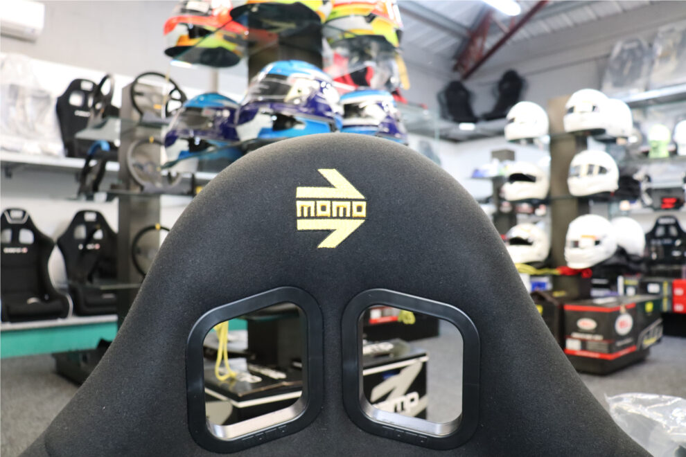 The top of a black MOMO bucket seat with a yellow MOMO logo and two harness slots, with racing helmets on display behind