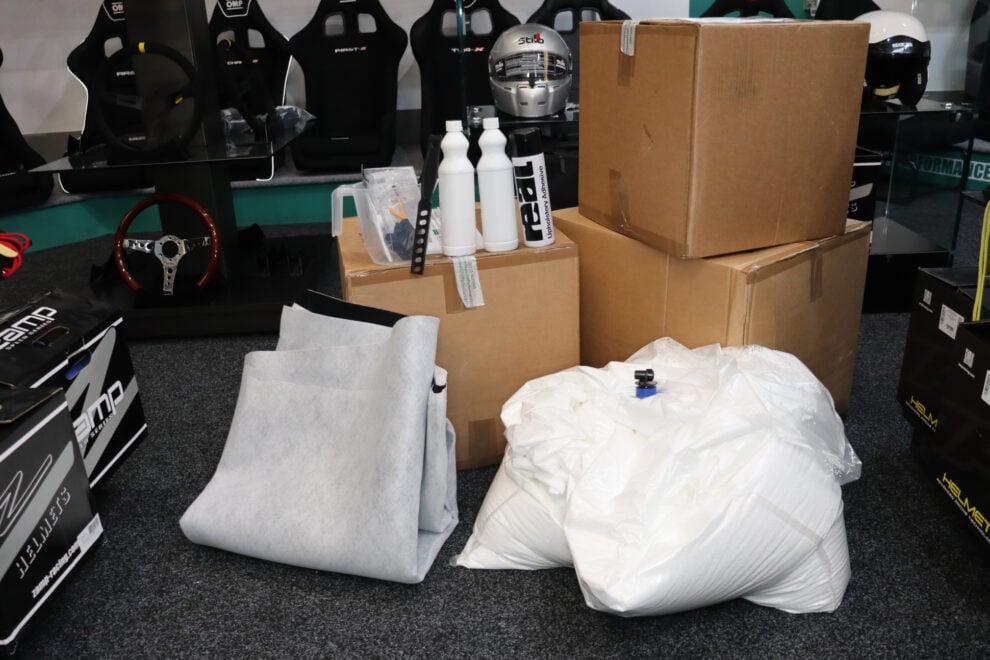 An unpacked seat insert kit on the showroom floor: a folded grey liner, a white bag of beads, bottles, a mixing jug and upholstery adhesive in front of boxes and racing seats