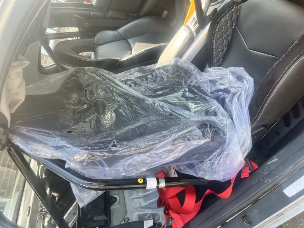 A black reclining sports seat still in its protective plastic, test fitted inside a car with a roll cage, its back reaching the roof lining