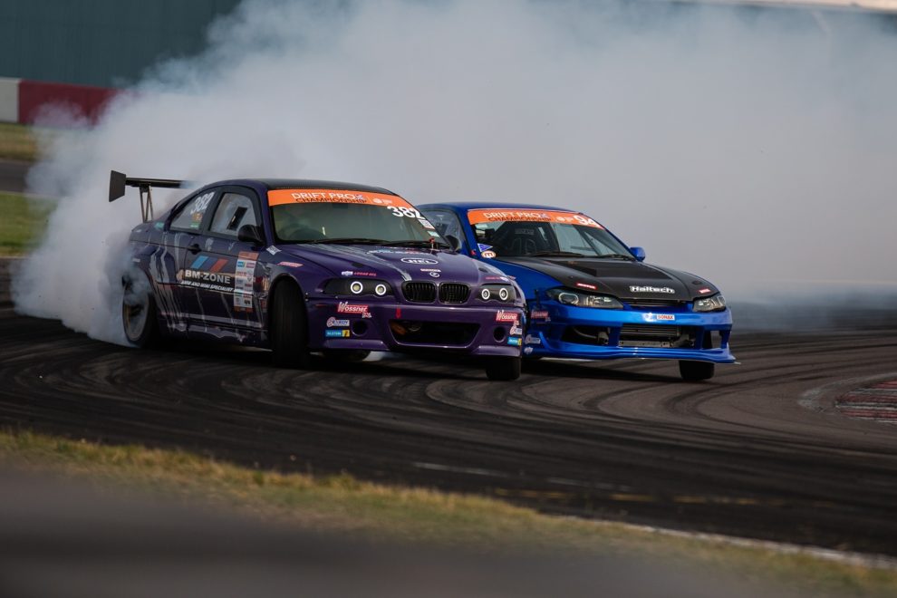 A purple BMW drift car chasing a blue Nissan Silvia through a corner in tyre smoke