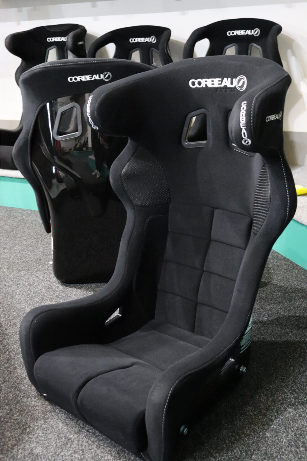 A black Corbeau Revenge bucket seat on the showroom floor, with the glossy shell of another seat and a row of Corbeau seats on the plinth behind