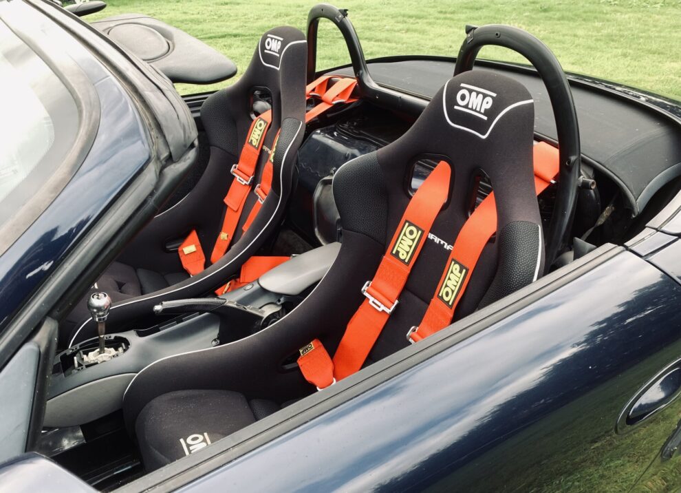 Two black OMP bucket seats with red OMP harnesses in a dark blue Porsche 986 Boxster, black hoops behind the seats