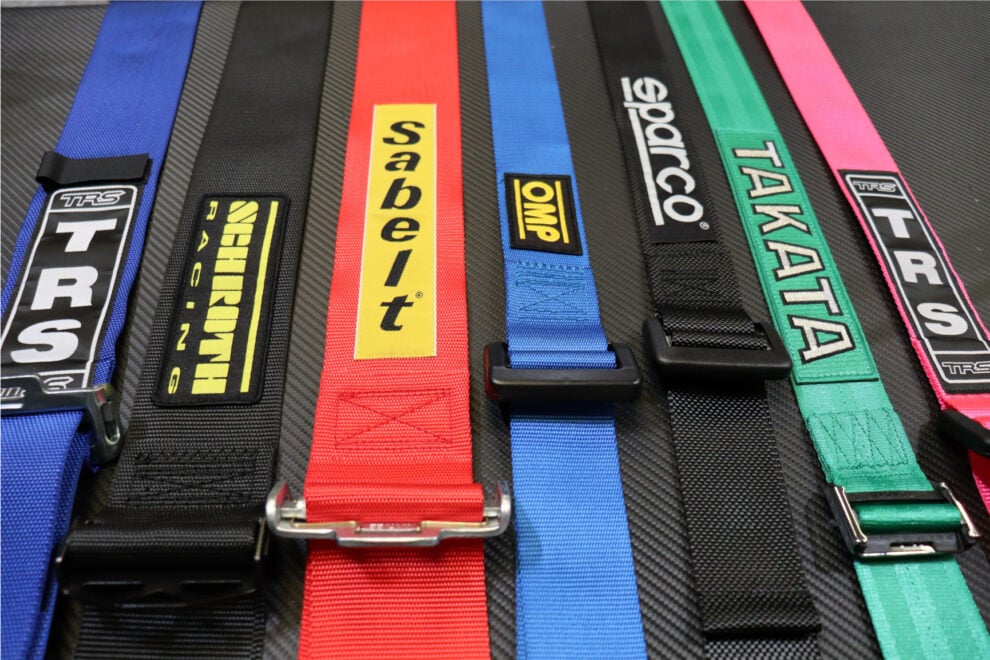 Harness straps from TRS, Schroth, Sabelt, OMP, Sparco and Takata laid side by side on a carbon fibre surface