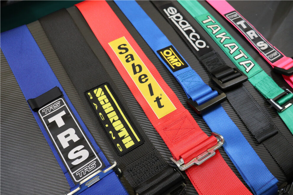 Harness straps from TRS, Schroth, Sabelt, OMP, Sparco and Takata laid side by side on a carbon effect surface