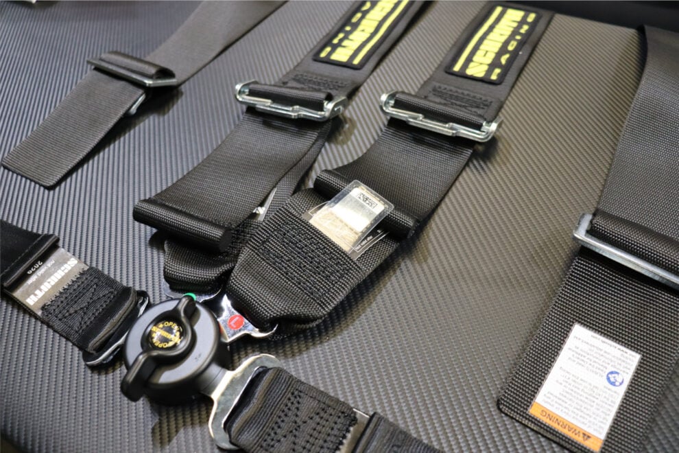 Black Schroth harness straps laid on a carbon surface, showing the metal webbing adjusters and the rotary buckle