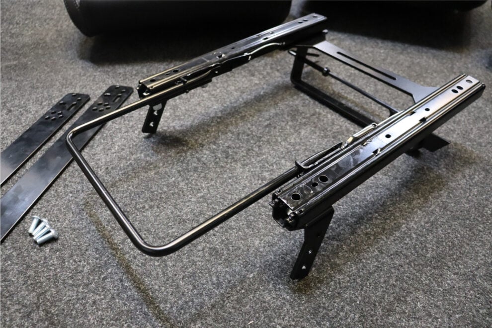 A Cobra Classic Mini subframe with its sliding runners on a carpeted floor, beside a pair of flat steel adaption bars and four bolts