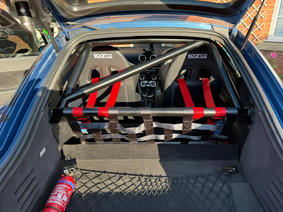 A black bolt-in rear roll cage with a diagonal and harness bar in the back of a customer's Audi TT, with red harness straps wrapped around the bar