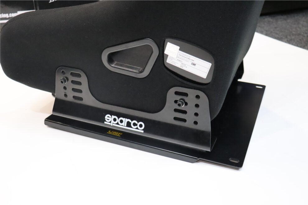 The side of a black OMP TRS-E XL bucket seat bolted to black Sparco side mounts on a base plate, with the FIA label visible