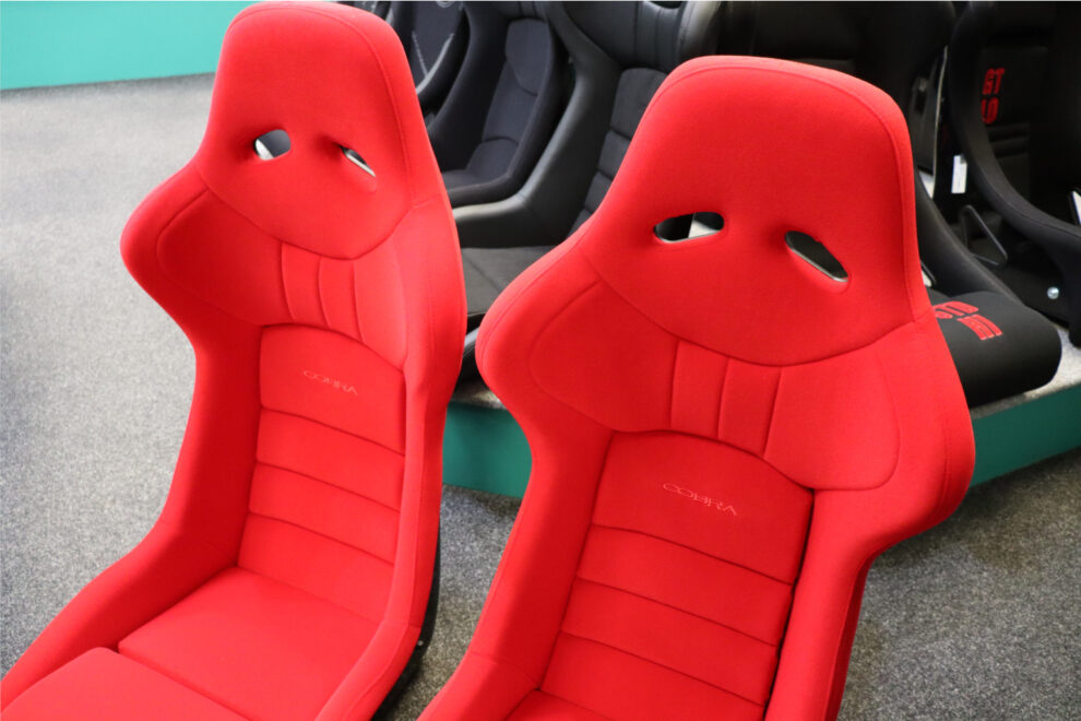 Two red velour Cobra Nogaro Club Sport Ebisu Edition seats on the showroom floor, with black seats on a display plinth behind