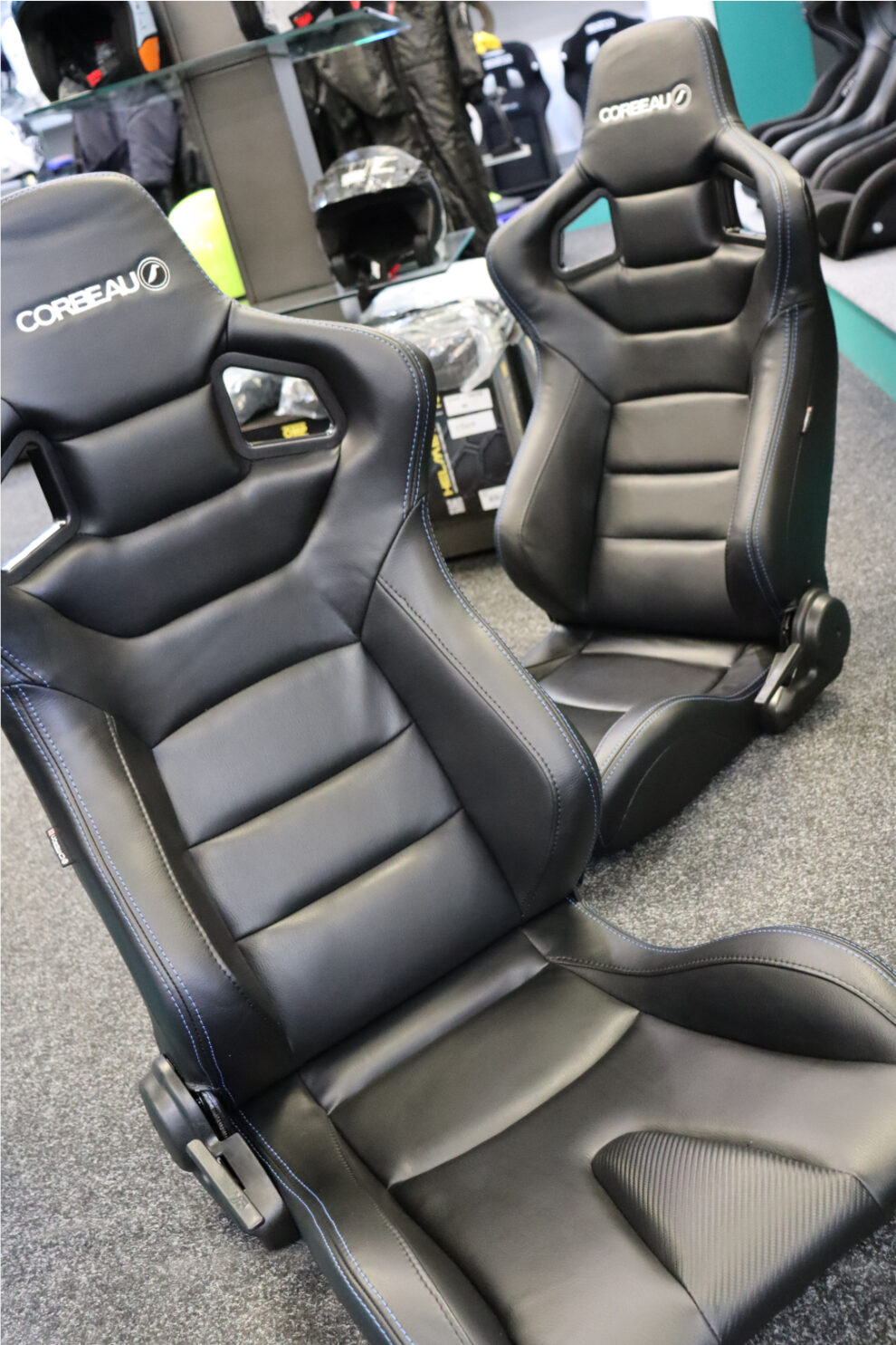 Two black Corbeau RRS seats with blue stitching seen at an angle in the showroom, with helmets and race suits behind