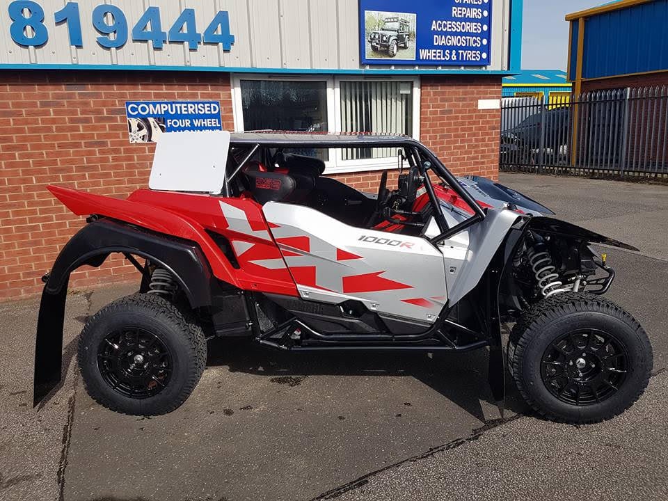 Side view of a red, silver and black Yamaha YXZ 1000R with red trimmed Mirco RS2 bucket seats, parked outside a 4x4 garage