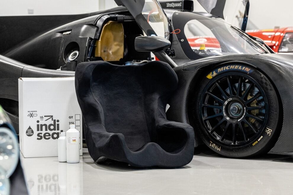 A finished black moulded indi seat insert standing beside its kit box and resin bottles, in front of a carbon prototype race car