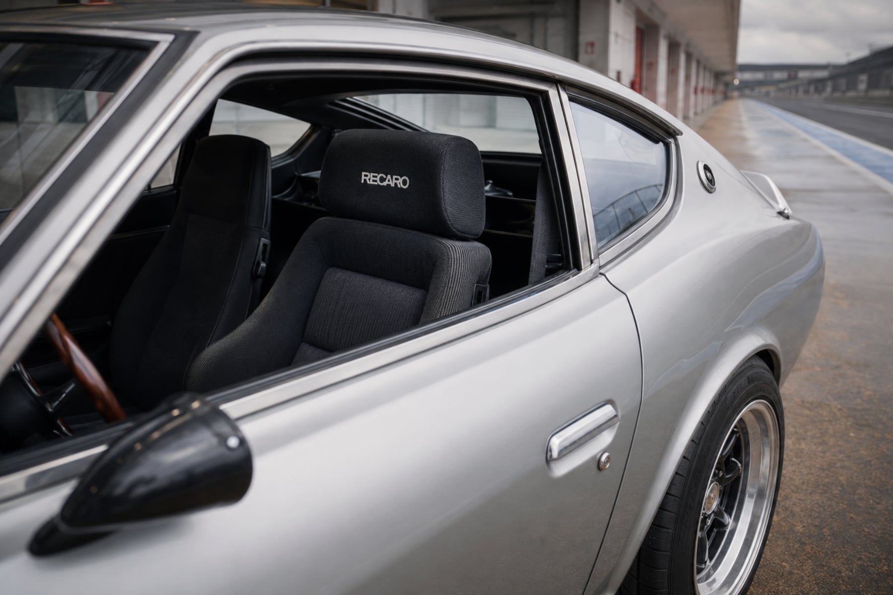 A silver Datsun 240Z S30 parked in a pit lane with the window down, showing a black Recaro reclining seat fitted inside, the Recaro name on the headrest