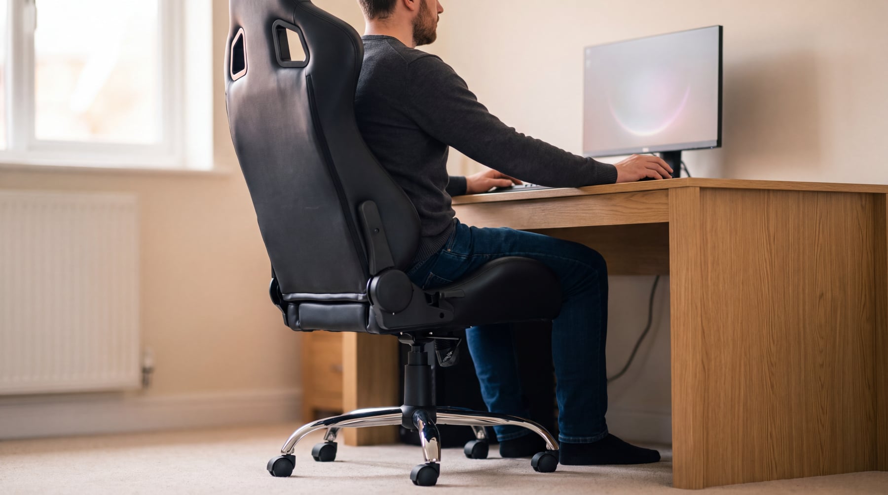 A man working at a desk in a black bucket sports seat converted into an office chair, with the chrome five arm base, castors and gas lift column in the foreground
