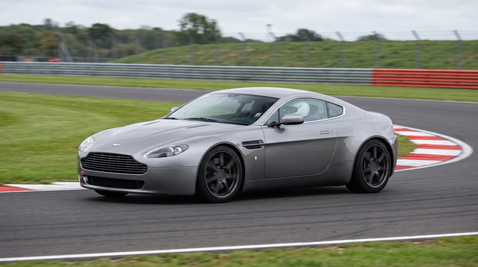 A silver 2007 Aston Martin V8 Vantage coupe with a helmeted driver cornering on the tarmac of a UK race circuit