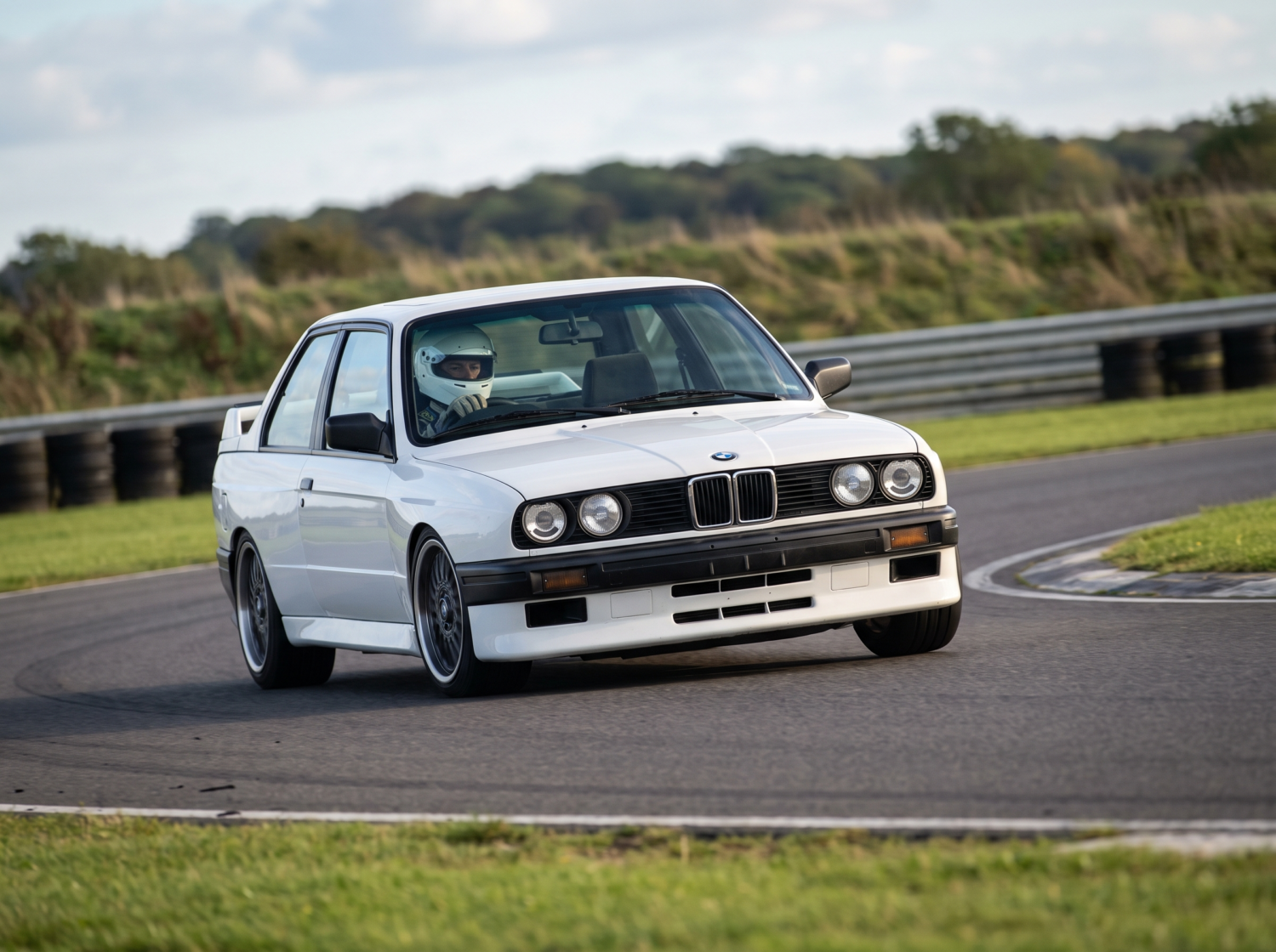White BMW M3 E30 cornering on a British race circuit, helmeted driver at the wheel