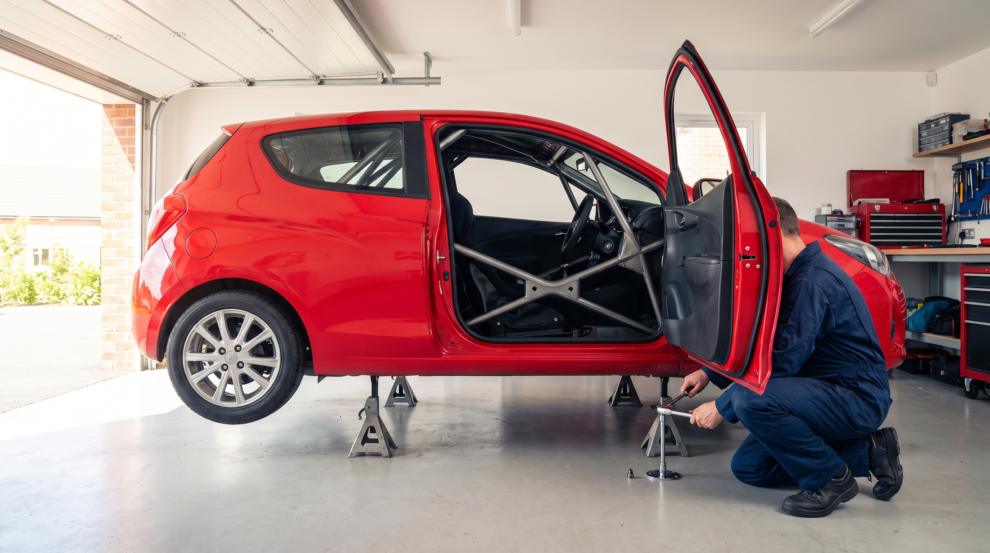 A red hatchback on axle stands in a home garage with a bolt-in roll cage visible through the open door, a mechanic tightening the mounting bolts