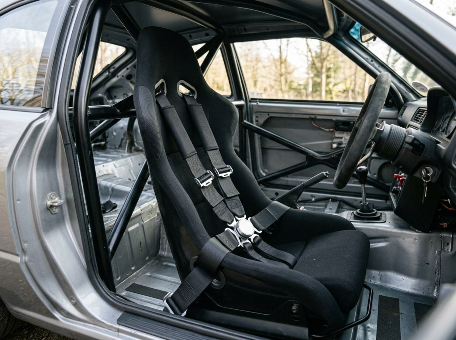 Six point racing harness with an alloy rotary buckle fastened across a black bucket seat in a caged race car