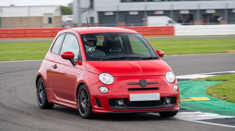 A red Fiat 595 Abarth cornering on a British race circuit with a helmeted driver behind the wheel