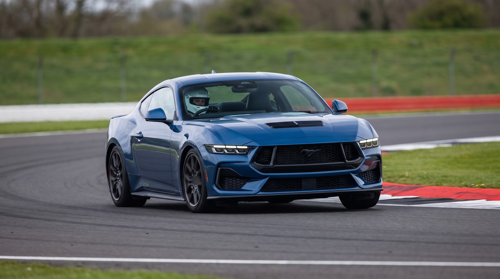 A blue 2024 Ford Mustang GT S650 with a helmeted driver cornering on the tarmac of a UK race circuit