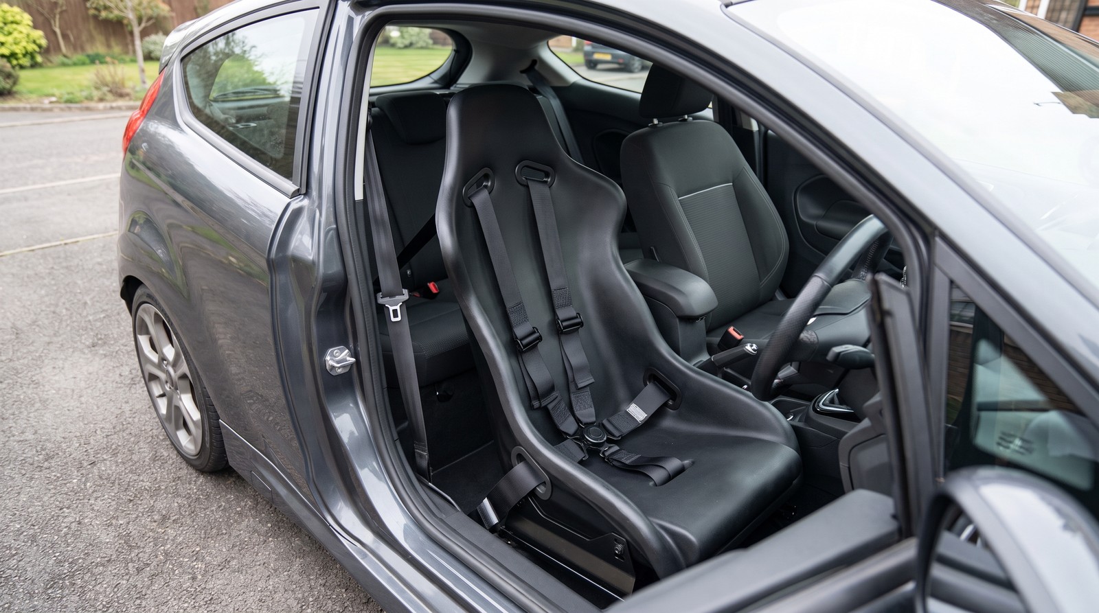 Plain black four point harness fitted to a bucket seat in a road hatchback with the standard rear bench and no roll cage, original seatbelt still on the pillar