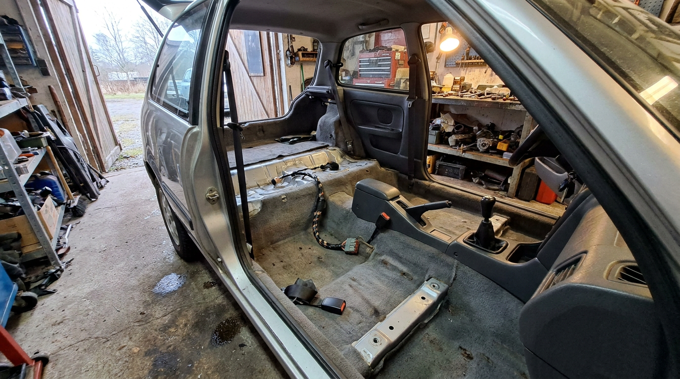 Interior of a three door hatchback with both front seats removed, showing the bare floor mounting points, a taped wiring connector and a loose seat belt stalk