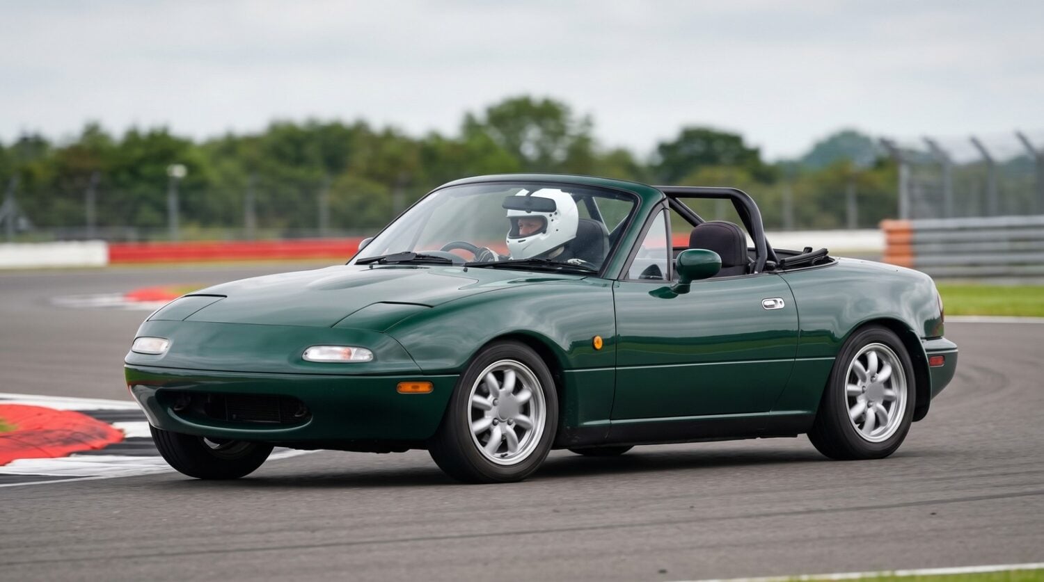 A British Racing Green Mazda MX-5 Mk1 with a helmeted driver cornering on a UK race circuit