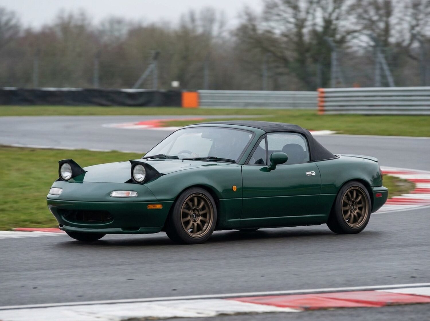 British racing green Mazda MX-5 Mk1 with pop-up headlights raised, cornering on a British race circuit