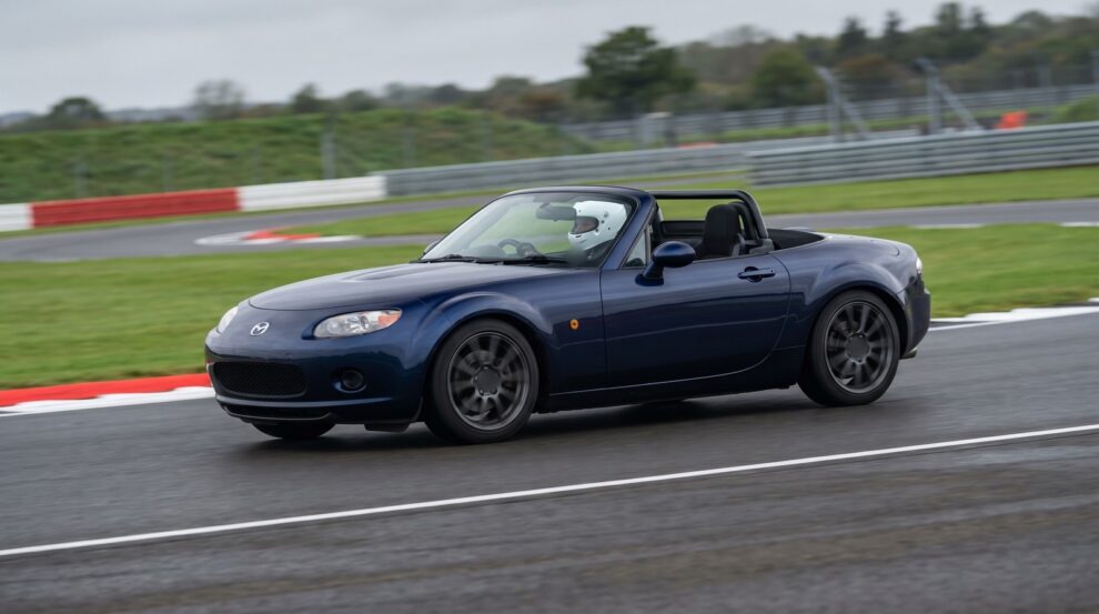 A dark blue Mazda MX-5 Mk3 NC with the roof down and a helmeted driver cornering on a UK race circuit