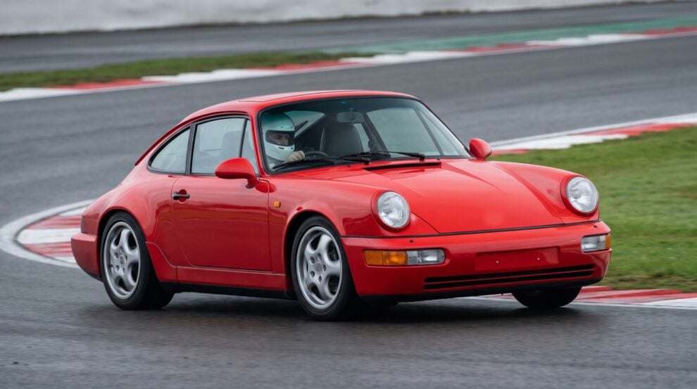 Guards red Porsche 911 964 cornering at an apex on a British race circuit with a helmeted driver at the wheel