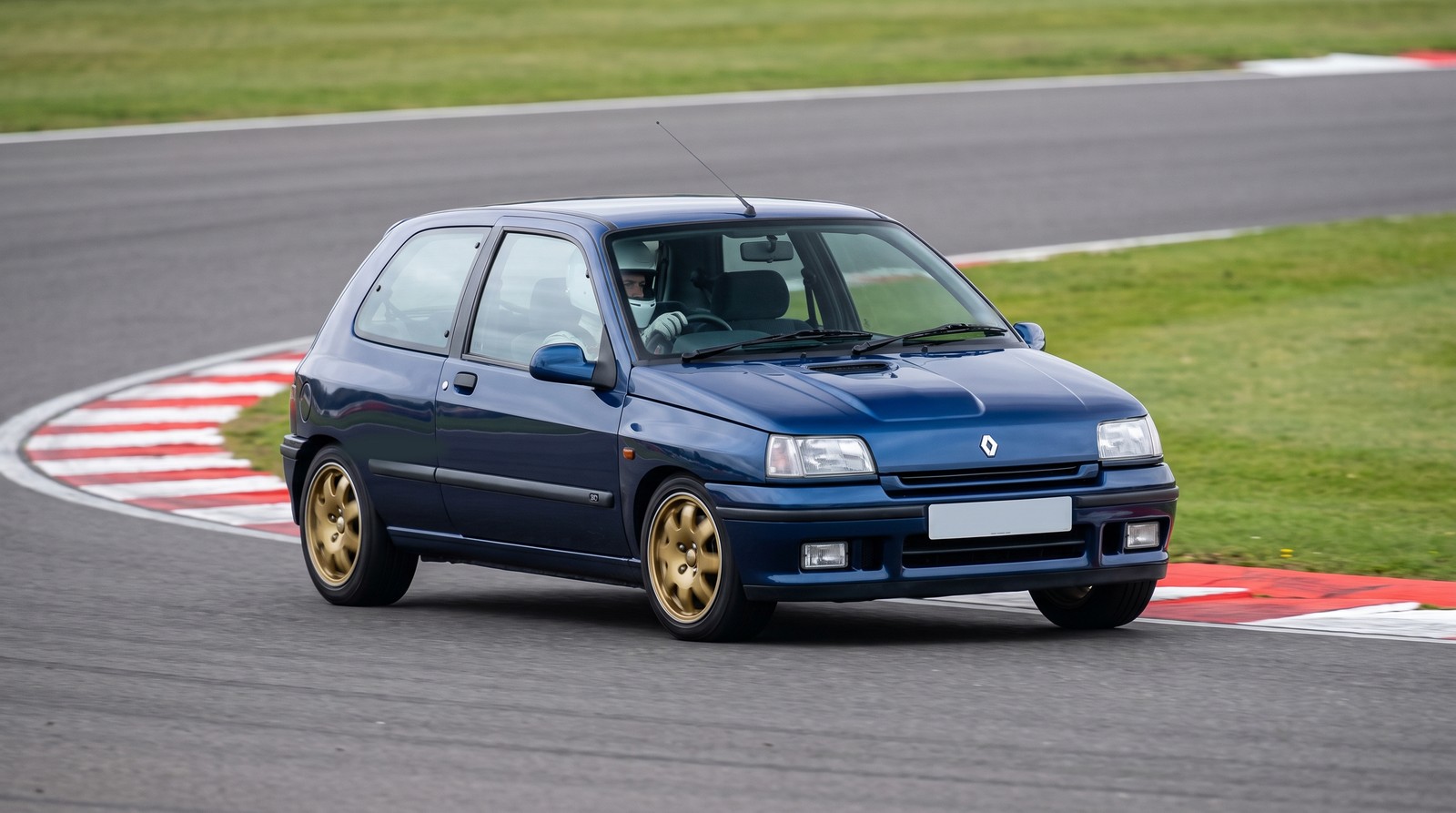 Blue Renault Clio Williams Mk1 hot hatch with gold alloys cornering at an apex on a British race circuit with a helmeted driver at the wheel