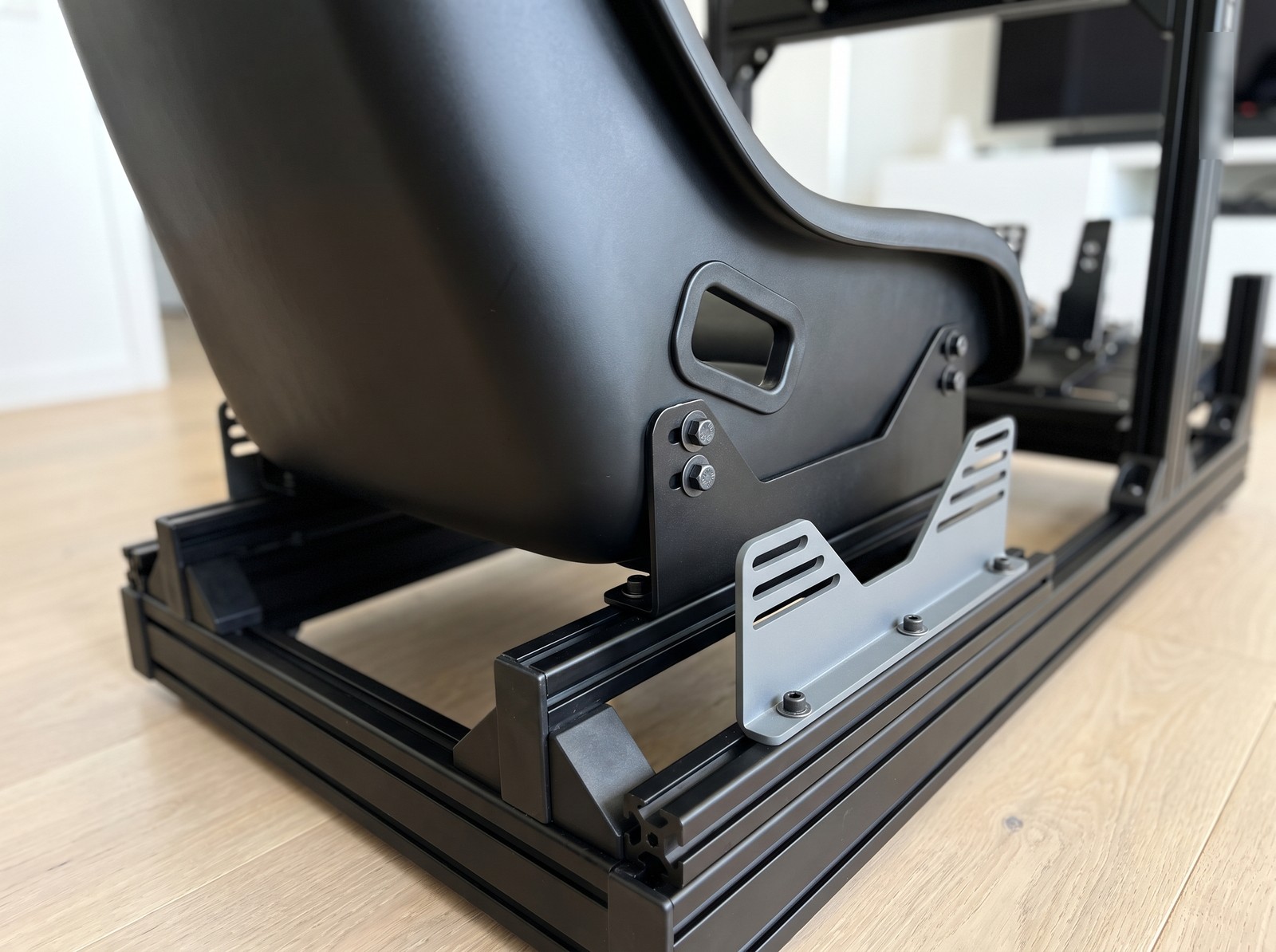 Plain black bucket seat on side mount brackets bolted to the matte black profile seat rails of a Sim-Lab style rig, grey slotted bracket alongside