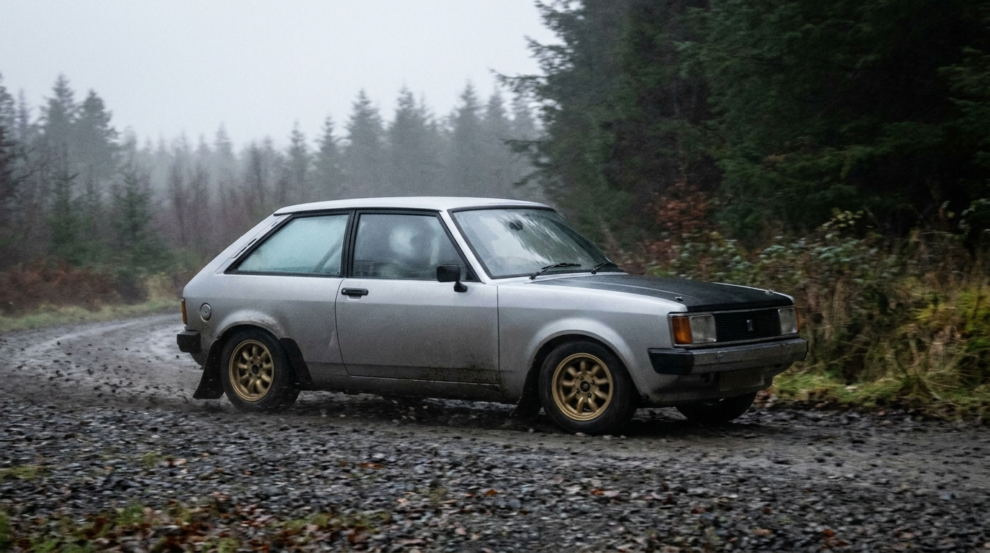 A silver Talbot Sunbeam three door with a black bonnet on a damp gravel forest rally stage