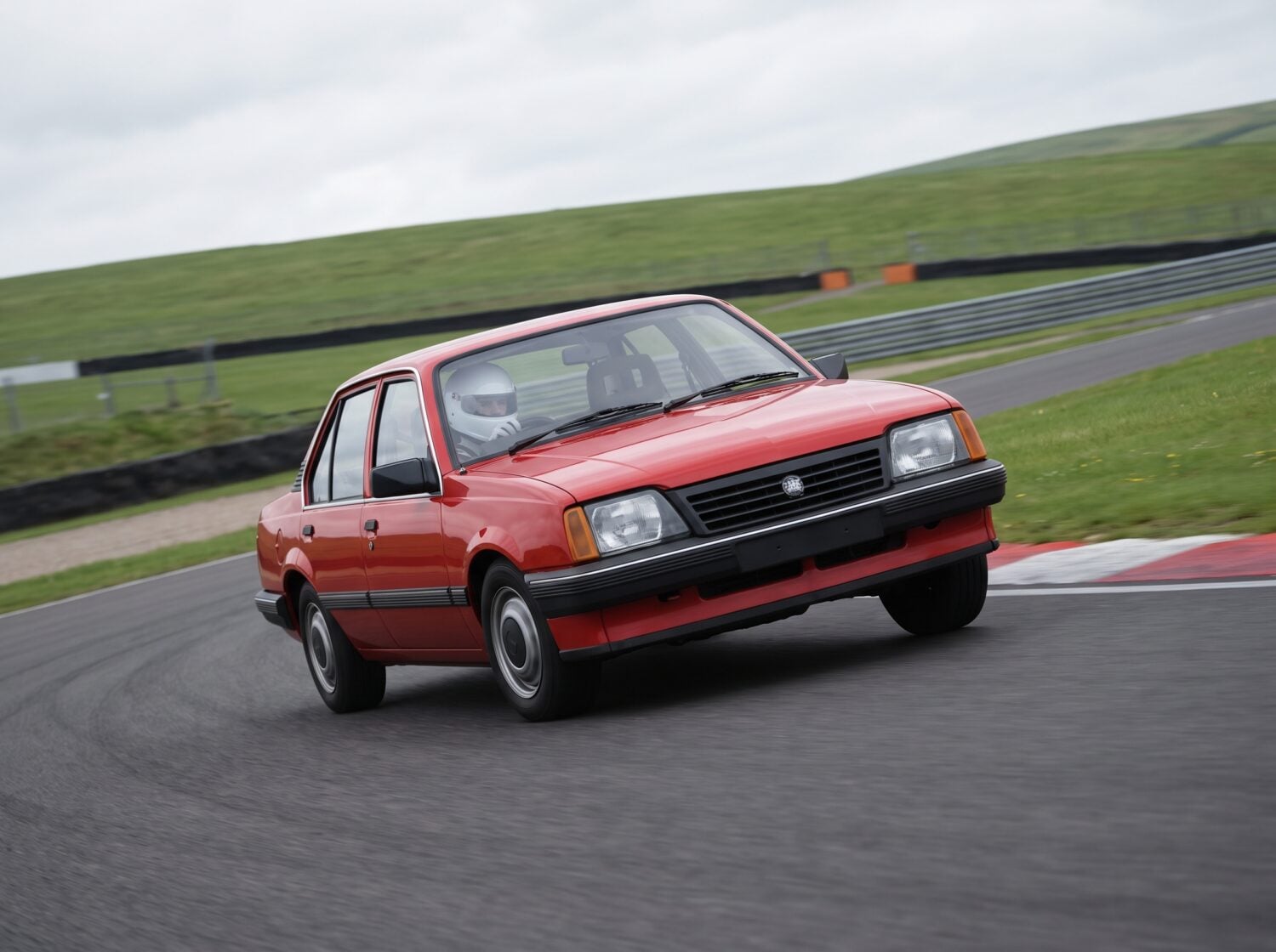 Red Vauxhall Cavalier Mk2 saloon cornering on a British race circuit, helmeted driver visible through the windscreen
