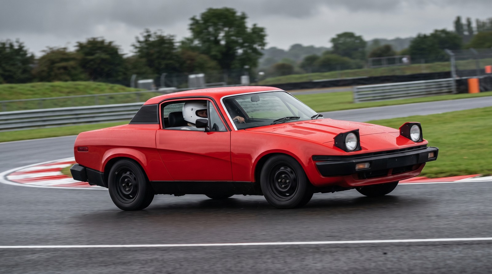 A red Triumph TR7 coupe with a helmeted driver cornering on the tarmac of a UK race circuit