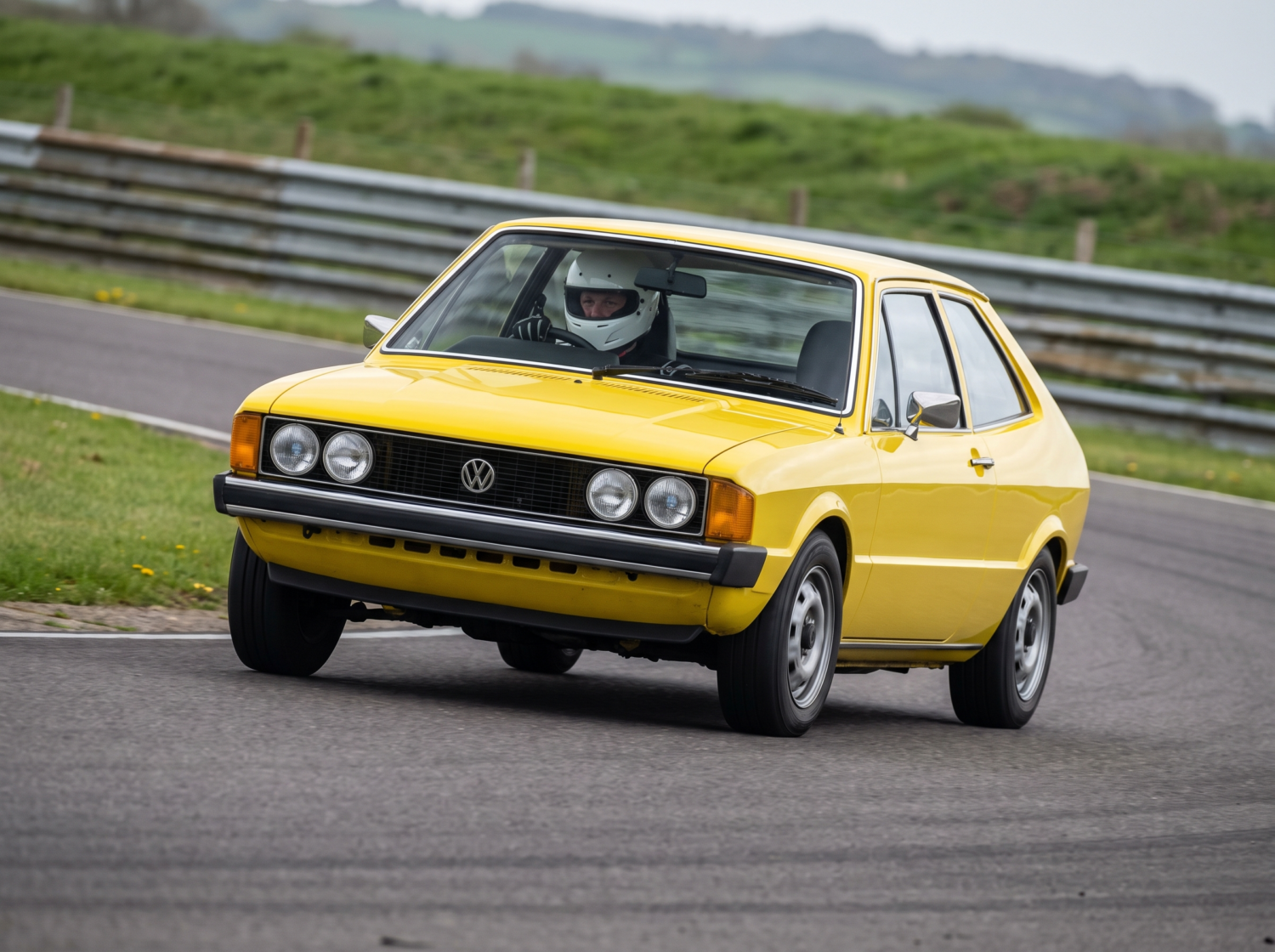 Yellow Volkswagen Scirocco Mk1 cornering on a British race circuit, helmeted driver at the wheel