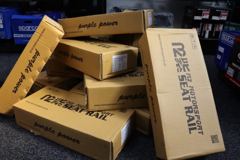 A pile of M2 Motorsport seat rail boxes in the GSM Performance showroom, the printed labels showing one set per box with left and right hand rails listed separately
